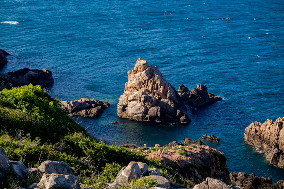 The image depicts a rocky coastline with a large, light brown rock formation jutting out into the deep blue ocean. The rock formation is surrounded by smaller rocks and boulders scattered across the shoreline. The water surrounding the rock formation appears calm and serene, reflecting the clear sky above.