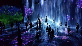 A group of people are standing and walking inside an immersive art installation with flowing digital streams and vibrant floral projections on the walls and floor. The environment appears interactive, with the participants seemingly engaged with the digital elements around them.