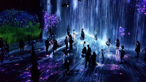 A group of people are standing and walking inside an immersive art installation with flowing digital streams and vibrant floral projections on the walls and floor. The environment appears interactive, with the participants seemingly engaged with the digital elements around them.