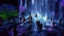 A group of people are standing and walking inside an immersive art installation with flowing digital streams and vibrant floral projections on the walls and floor. The environment appears interactive, with the participants seemingly engaged with the digital elements around them.
