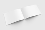 An open, blank booklet with white pages on a light gray background, displayed in a slightly tilted position.