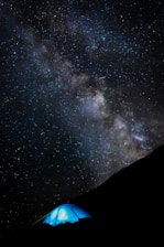 A vast, star-filled night sky with the Milky Way galaxy prominently visible, arching overhead. Below, a glowing blue tent is set up on a darkened hillside, suggesting a camping scene. The contrast between the bright stars and the darkness of the landscape is striking.