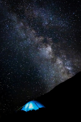 A vast, star-filled night sky with the Milky Way galaxy prominently visible, arching overhead. Below, a glowing blue tent is set up on a darkened hillside, suggesting a camping scene. The contrast between the bright stars and the darkness of the landscape is striking.
