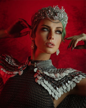 A person wearing an ornate silver crown adorned with intricate designs and decorations stands out against a deep red background. The individual has green hair and is dressed in a shimmering chainmail outfit with layered metallic scales, giving a regal and warrior-like appearance.