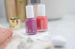 Two bottles of nail polish in pink and coral shades are placed on a light-colored surface. They are surrounded by soft-focus objects such as a gold cylindrical container and a folded pink towel, creating a cozy and elegant ambience.