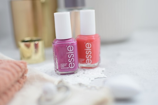 Two bottles of nail polish in pink and coral shades are placed on a light-colored surface. They are surrounded by soft-focus objects such as a gold cylindrical container and a folded pink towel, creating a cozy and elegant ambience.