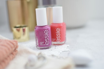 Two bottles of nail polish in pink and coral shades are placed on a light-colored surface. They are surrounded by soft-focus objects such as a gold cylindrical container and a folded pink towel, creating a cozy and elegant ambience.
