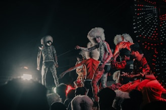 Performers dressed in tiger-like costumes with white furry headpieces engage in a vibrant street parade or performance, surrounded by a crowd. The scene is filled with colorful lights and dynamic movement.