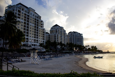 Tall residential or hotel buildings line a sandy beach, with lounge chairs and a lifeguard stand visible. Palm trees are scattered throughout, and the ocean is calm under a cloudy sunset sky. A small boat is docked near the shore.
