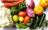 An assortment of fresh vegetables and fruits including carrots, bell peppers, turnips, two oranges, cabbage, tomatoes, cucumbers, green chili peppers, and some mint leaves arranged on a white background.