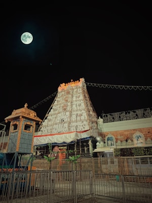 A night scene featuring an ornately carved temple gopuram illuminated against the night sky with a full moon. The gopuram is adorned with intricate sculptures and surrounded by a gold-colored structure. Decorative lights are strung across, enhancing the majestic appearance.