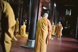 A group of people dressed in orange robes are standing and kneeling in a dimly lit interior space. The setting appears to be a temple or religious hall, with wooden columns and red tiled flooring. Some individuals have their hands in prayer position, creating a serene and contemplative atmosphere.