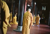 A group of people dressed in orange robes are standing and kneeling in a dimly lit interior space. The setting appears to be a temple or religious hall, with wooden columns and red tiled flooring. Some individuals have their hands in prayer position, creating a serene and contemplative atmosphere.