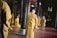 A group of people dressed in orange robes are standing and kneeling in a dimly lit interior space. The setting appears to be a temple or religious hall, with wooden columns and red tiled flooring. Some individuals have their hands in prayer position, creating a serene and contemplative atmosphere.