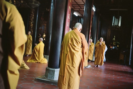 A group of people dressed in orange robes are standing and kneeling in a dimly lit interior space. The setting appears to be a temple or religious hall, with wooden columns and red tiled flooring. Some individuals have their hands in prayer position, creating a serene and contemplative atmosphere.