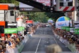 A street race with cyclists moving towards the camera as spectators line both sides of the road, captured during a cycling event. Large sponsor logos like ŠKODA are prominent, and there are several colorful balloons and advertising banners. A crowd of onlookers is focused on the action, capturing photographs and cheering.