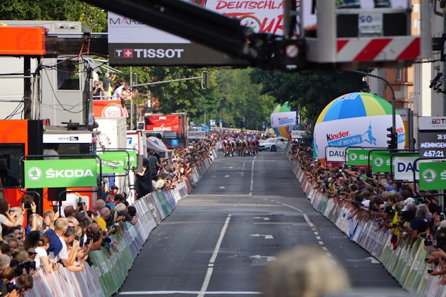 A street race with cyclists moving towards the camera as spectators line both sides of the road, captured during a cycling event. Large sponsor logos like ŠKODA are prominent, and there are several colorful balloons and advertising banners. A crowd of onlookers is focused on the action, capturing photographs and cheering.