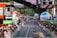 A street race with cyclists moving towards the camera as spectators line both sides of the road, captured during a cycling event. Large sponsor logos like ŠKODA are prominent, and there are several colorful balloons and advertising banners. A crowd of onlookers is focused on the action, capturing photographs and cheering.