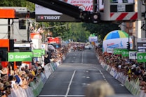 A street race with cyclists moving towards the camera as spectators line both sides of the road, captured during a cycling event. Large sponsor logos like &Scaron;KODA are prominent, and there are several colorful balloons and advertising banners. A crowd of onlookers is focused on the action, capturing photographs and cheering.