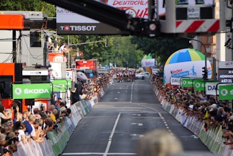 A street race with cyclists moving towards the camera as spectators line both sides of the road, captured during a cycling event. Large sponsor logos like ŠKODA are prominent, and there are several colorful balloons and advertising banners. A crowd of onlookers is focused on the action, capturing photographs and cheering.