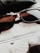 A pair of tortoiseshell sunglasses rests on a fluffy white surface. A black leather accessory is partially visible in the background, while there is a portion of a monochrome magazine or photo underneath.