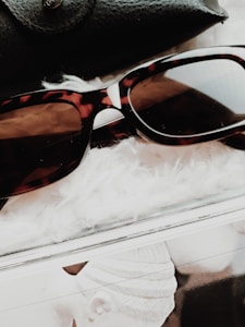 A pair of tortoiseshell sunglasses rests on a fluffy white surface. A black leather accessory is partially visible in the background, while there is a portion of a monochrome magazine or photo underneath.