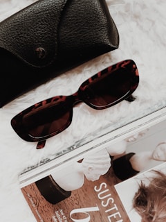 A pair of stylish tortoiseshell sunglasses is laid next to a black leather glasses case and a magazine on a soft, white surface. The magazine features a monochrome image and text.