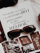 An open magazine displays a page with stylish typography in Portuguese and has a focus on fashion accessories such as sunglasses and jewelry. The sunglasses rest on top of the open page, with additional images showcasing intricate jewelry pieces.