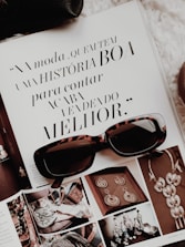 An open magazine displays a page with stylish typography in Portuguese and has a focus on fashion accessories such as sunglasses and jewelry. The sunglasses rest on top of the open page, with additional images showcasing intricate jewelry pieces.