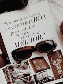 An open magazine displays a page with stylish typography in Portuguese and has a focus on fashion accessories such as sunglasses and jewelry. The sunglasses rest on top of the open page, with additional images showcasing intricate jewelry pieces.