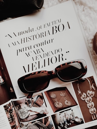 An open magazine displays a page with stylish typography in Portuguese and has a focus on fashion accessories such as sunglasses and jewelry. The sunglasses rest on top of the open page, with additional images showcasing intricate jewelry pieces.