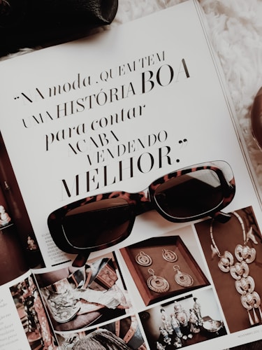 An open magazine displays a page with stylish typography in Portuguese and has a focus on fashion accessories such as sunglasses and jewelry. The sunglasses rest on top of the open page, with additional images showcasing intricate jewelry pieces.