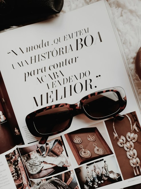 An open magazine displays a page with stylish typography in Portuguese and has a focus on fashion accessories such as sunglasses and jewelry. The sunglasses rest on top of the open page, with additional images showcasing intricate jewelry pieces.