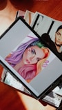 A digital tablet displaying an illustration of a woman with vibrant rainbow-colored hair, featuring shades of purple, green, yellow, and red. The woman's makeup is bold with colorful eyeshadow and red lipstick. The tablet is resting on a stack of magazines, one of which has a black and white cover with a portrait of a person.