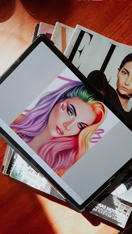 A digital tablet displaying an illustration of a woman with vibrant rainbow-colored hair, featuring shades of purple, green, yellow, and red. The woman's makeup is bold with colorful eyeshadow and red lipstick. The tablet is resting on a stack of magazines, one of which has a black and white cover with a portrait of a person.