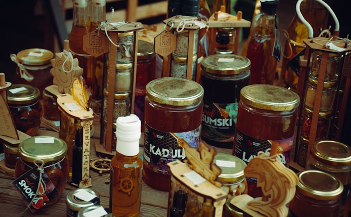A variety of jars and bottles displaying honey, dried fruits, and other artisanal products. The jars are arranged on a wooden surface, with decorative wooden signs and labels highlighting the contents. The products appear to be homemade or locally sourced, giving an artisanal and rustic vibe.