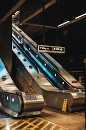 A modern indoor setting features an escalator in a metro or train station. The escalator appears to be metallic and illuminated, with a person standing on it. Overhead signs indicate directions such as 'Way out' and 'DLR', and a caution sign is placed on the ground. The environment has an industrial design with visible structures.