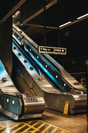 A modern indoor setting features an escalator in a metro or train station. The escalator appears to be metallic and illuminated, with a person standing on it. Overhead signs indicate directions such as 'Way out' and 'DLR', and a caution sign is placed on the ground. The environment has an industrial design with visible structures.