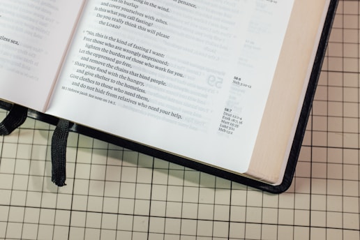 An open book on a grid-patterned surface displaying text with a black ribbon bookmark. The text appears to be scriptural or poetic in nature with verses and annotations.