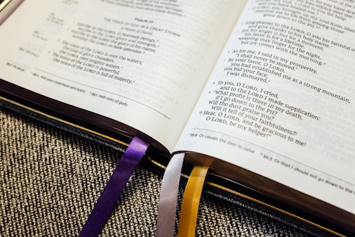 An open book with visible text, likely a religious scripture, rests on a textured fabric surface. The book has two colored ribbons, purple and yellow, marking different pages.