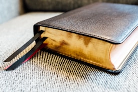 A large book with a dark leather cover and gold-edged pages rests on a textured fabric surface. Two ribbons, one black and one red, extend from the book's spine, serving as bookmarks.