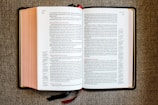 An open book with red and black text on white pages, displayed on a textured fabric surface. The book appears to be a religious or biblical text, with sections and chapter numbers in red and verse numbers in black. Two ribbon bookmarks hang from the bottom.