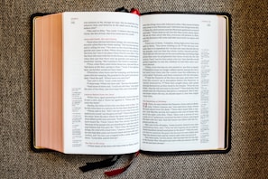 An open book with red and black text on white pages, displayed on a textured fabric surface. The book appears to be a religious or biblical text, with sections and chapter numbers in red and verse numbers in black. Two ribbon bookmarks hang from the bottom.