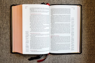 An open book with red and black text on white pages, displayed on a textured fabric surface. The book appears to be a religious or biblical text, with sections and chapter numbers in red and verse numbers in black. Two ribbon bookmarks hang from the bottom.