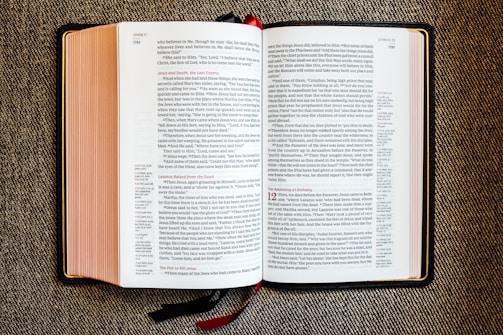 An open book with red and black text on white pages, displayed on a textured fabric surface. The book appears to be a religious or biblical text, with sections and chapter numbers in red and verse numbers in black. Two ribbon bookmarks hang from the bottom.