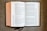 An open book with red and black text on white pages, displayed on a textured fabric surface. The book appears to be a religious or biblical text, with sections and chapter numbers in red and verse numbers in black. Two ribbon bookmarks hang from the bottom.