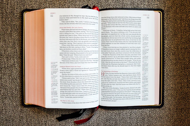 An open book with red and black text on white pages, displayed on a textured fabric surface. The book appears to be a religious or biblical text, with sections and chapter numbers in red and verse numbers in black. Two ribbon bookmarks hang from the bottom.