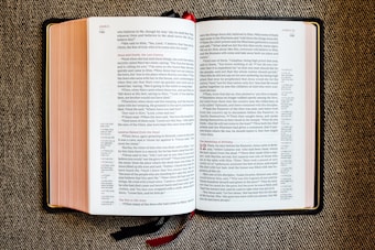 An open book with red and black text on white pages, displayed on a textured fabric surface. The book appears to be a religious or biblical text, with sections and chapter numbers in red and verse numbers in black. Two ribbon bookmarks hang from the bottom.