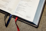 An open Bible is displayed on a fabric surface. Two ribbons, one blue and one red, act as bookmarks. The pages are filled with text, featuring a section labeled Psalm 142.