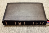 A black leather-bound book with the title 'Holy Bible' embossed in gold on the spine is lying flat on a textured surface. The book is thick, with several multi-colored ribbon markers peeking out from the bottom right corner.